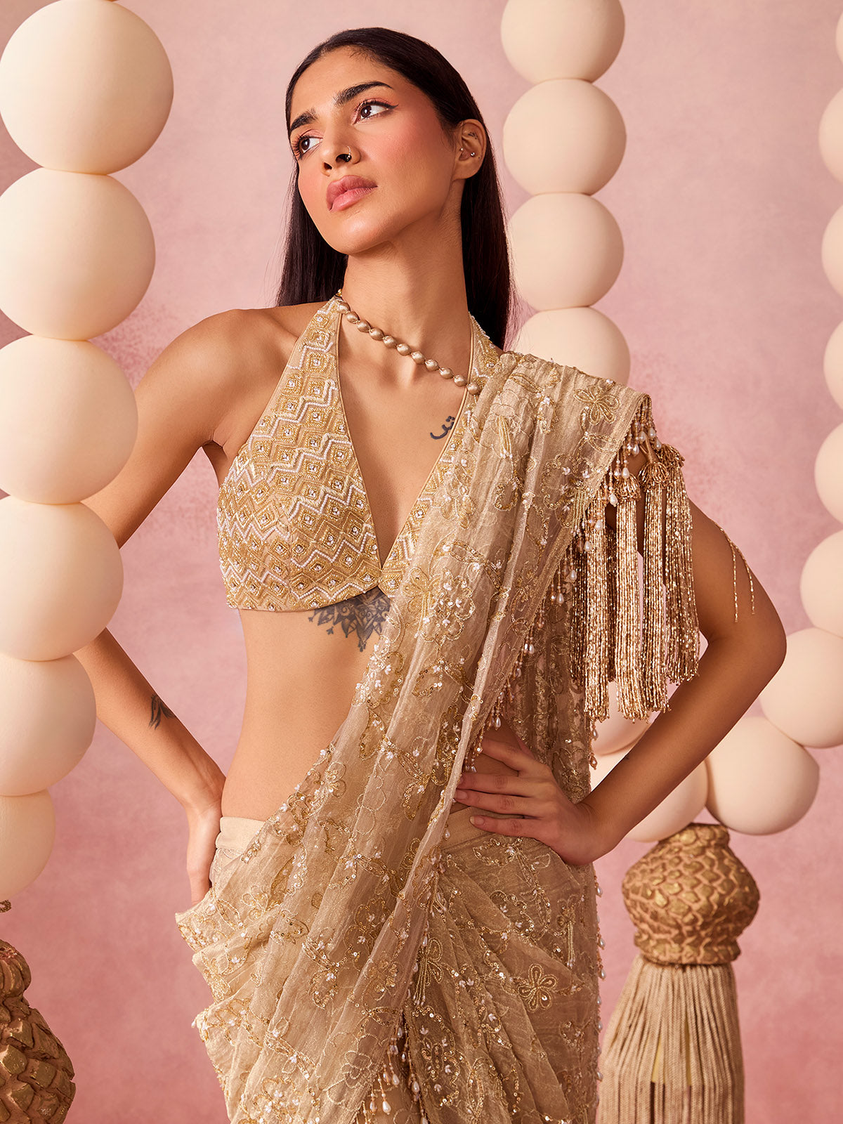 A woman standing in front of a pink background with large bead decor, wearing gold saree. 